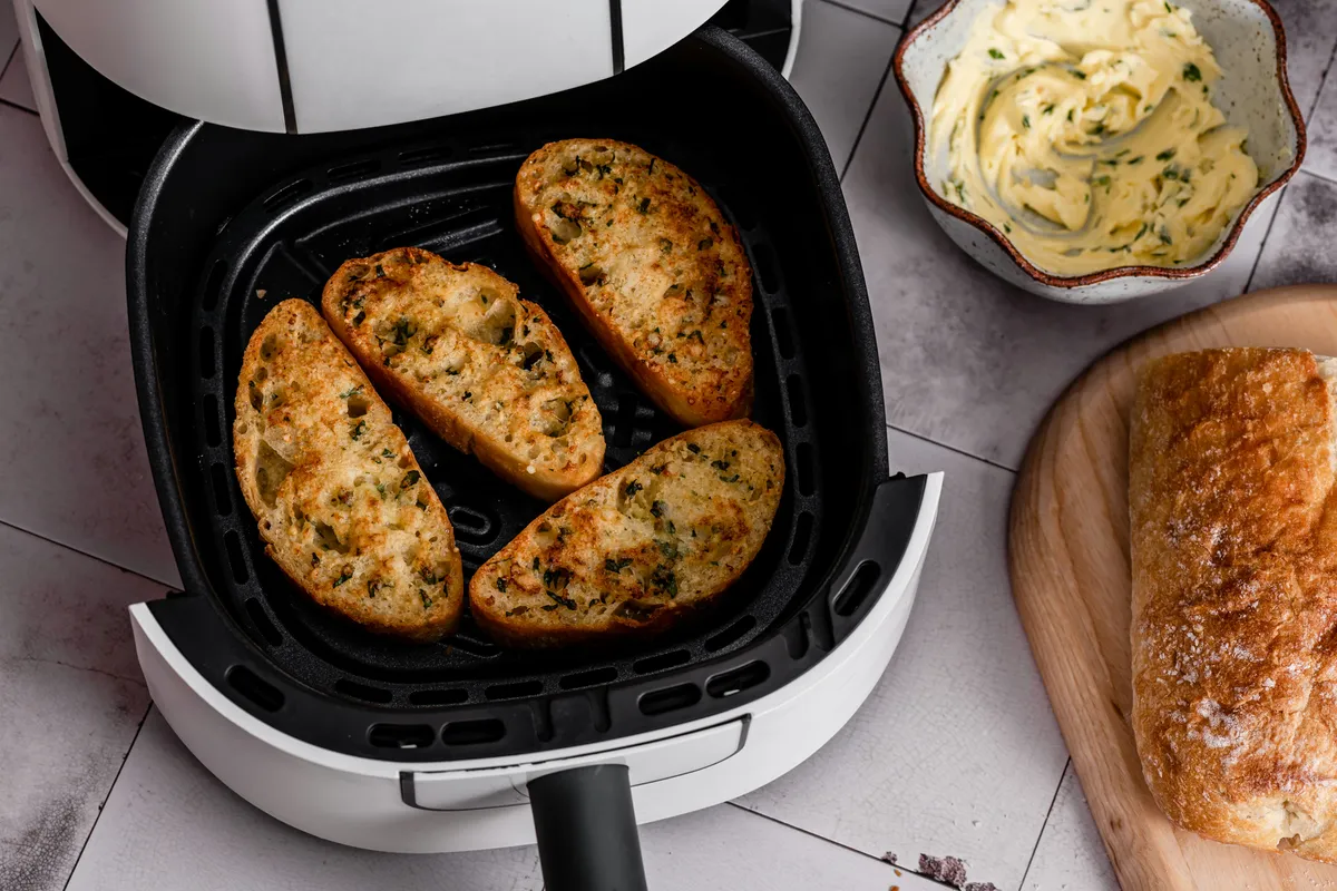 Slices of garlic bread browning in an air fryer basket next to a bowl of herb butter