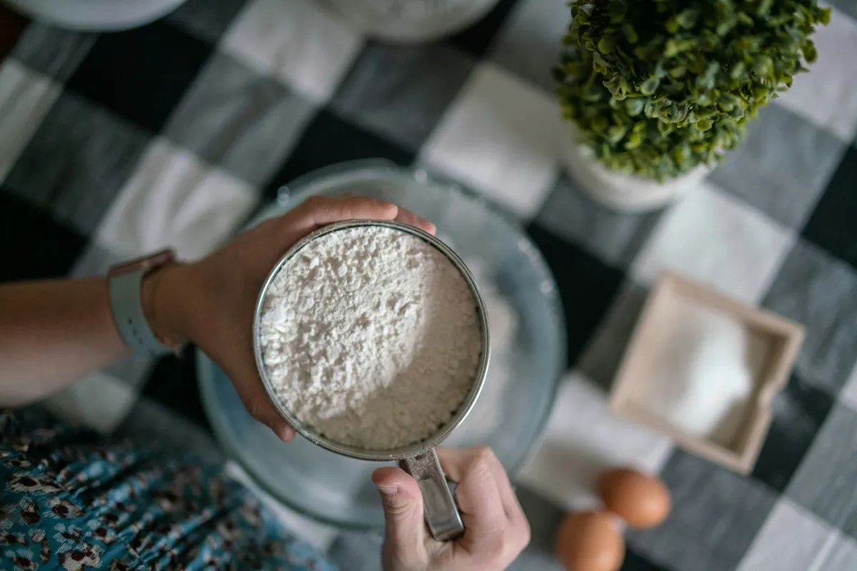 Hands holding a metal flour sifter over a glass mixing bowl with eggs nearby