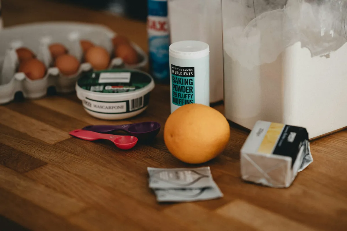 Baking powder tub, eggs, measuring spoons, butter and an orange on a wooden counter