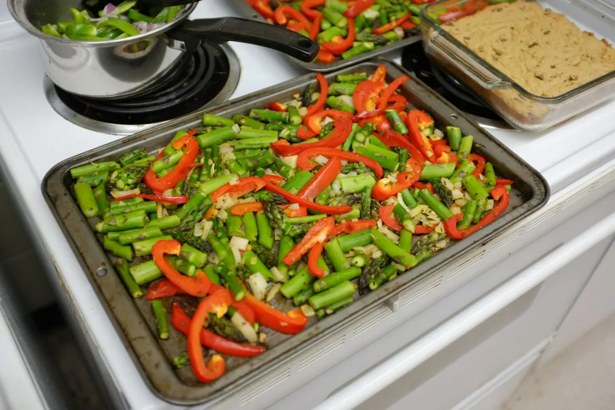 Sheet pans of cut red peppers, asparagus and onion on a white stove top