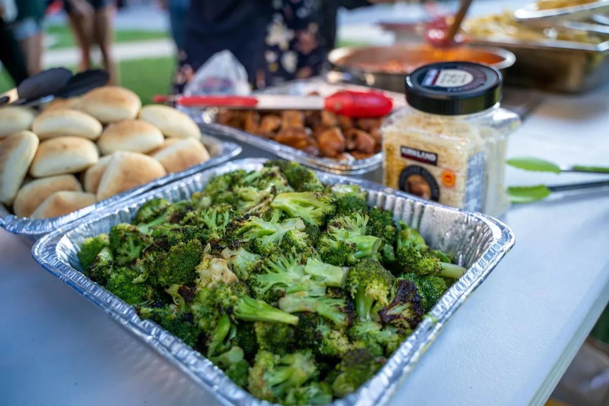 Charred roasted broccoli in a foil pan beside a tray of dinner rolls