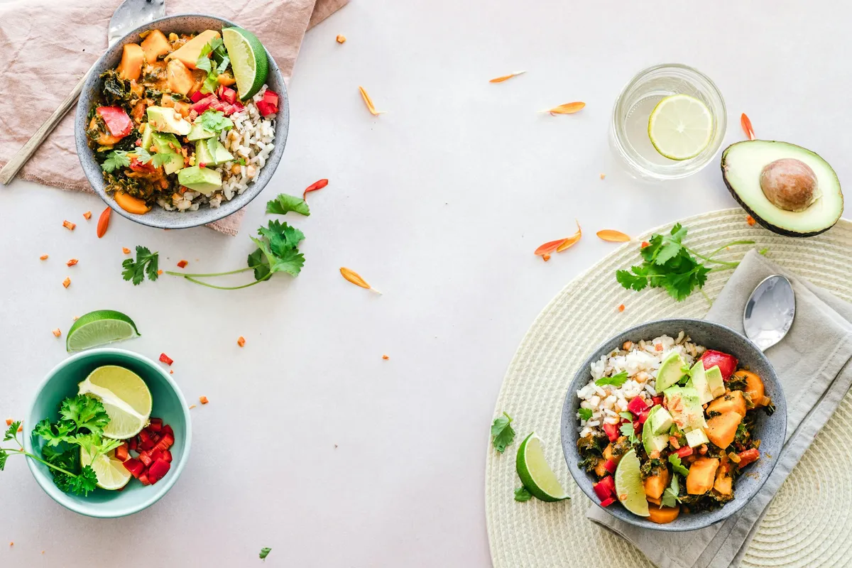 Two grain bowls with sweet potato, kale, avocado and red pepper beside a bowl of lime and parsley