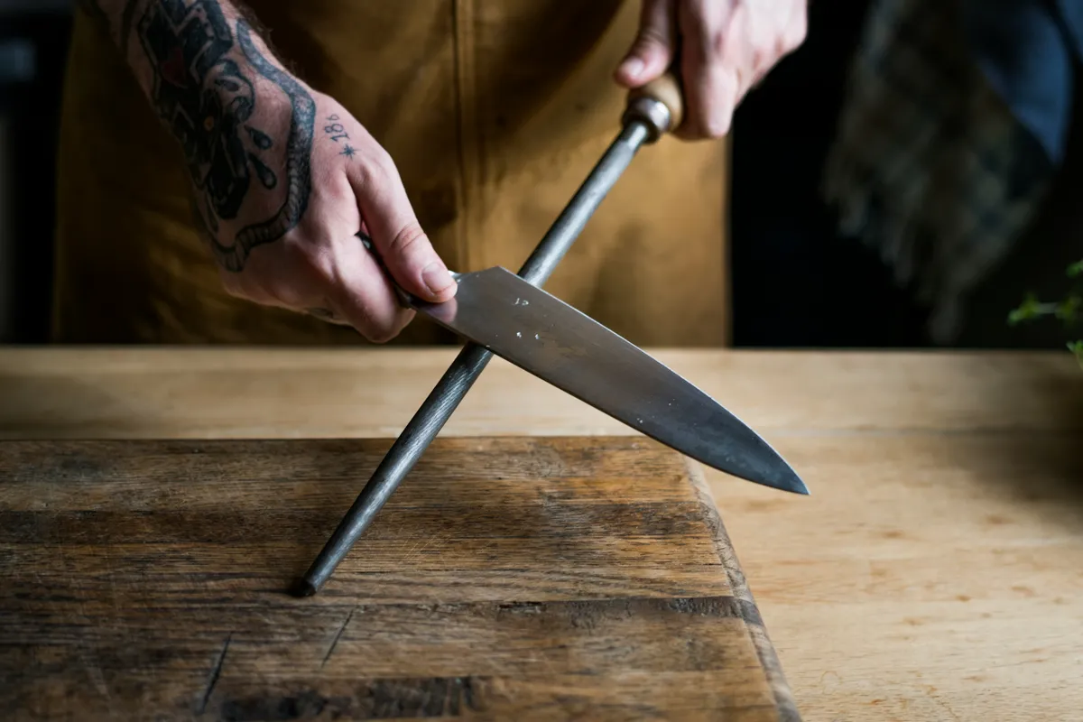 A cook running a chef knife along a honing steel over a wooden bench