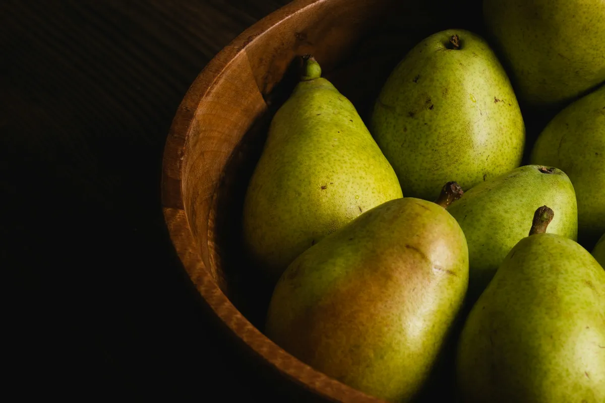 Green pears piled in a wooden bowl on a dark counter in low evening light